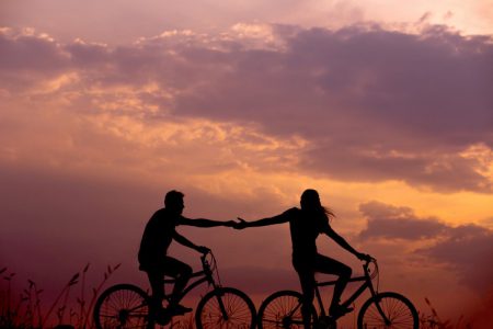 Två personer som cyklar i solnedgång, de håller varandra i handen som för att dra varandra framåt
