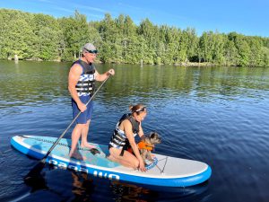 Två personer och en liten hund på en SUP-bräda i vattnet. I bakgrunden syns en skog. 