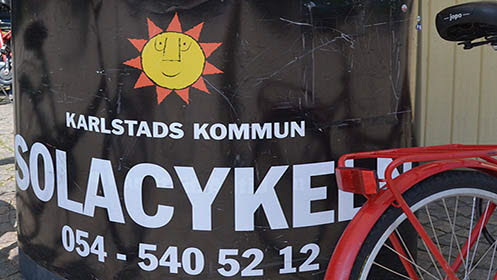 skylt med texten Solacykeln, Karlstads kommuns logga samt telefonnumret till Solacykeln. I högra hörnet syns delar av en röd cykel.