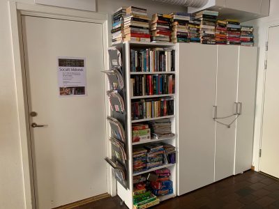 En bokhylla fylld med massa böcker men också spel. På en dörr sitter en affisch som beskriver Stadsmissionens sociala bibliotek.