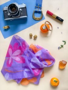 Detaljbild med en blommig lila scarf, blått skärp, kamera och accessoarer.